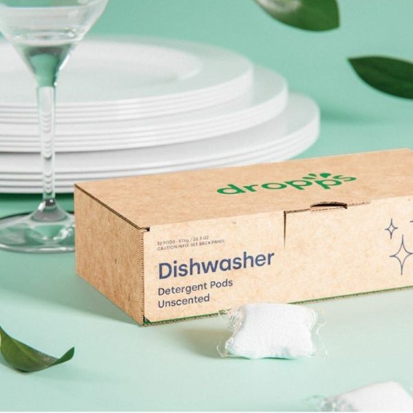 box of Dropps Dishwasher Pods and a couple of loose pods and plates and glass in the background on green table cloth with leaves